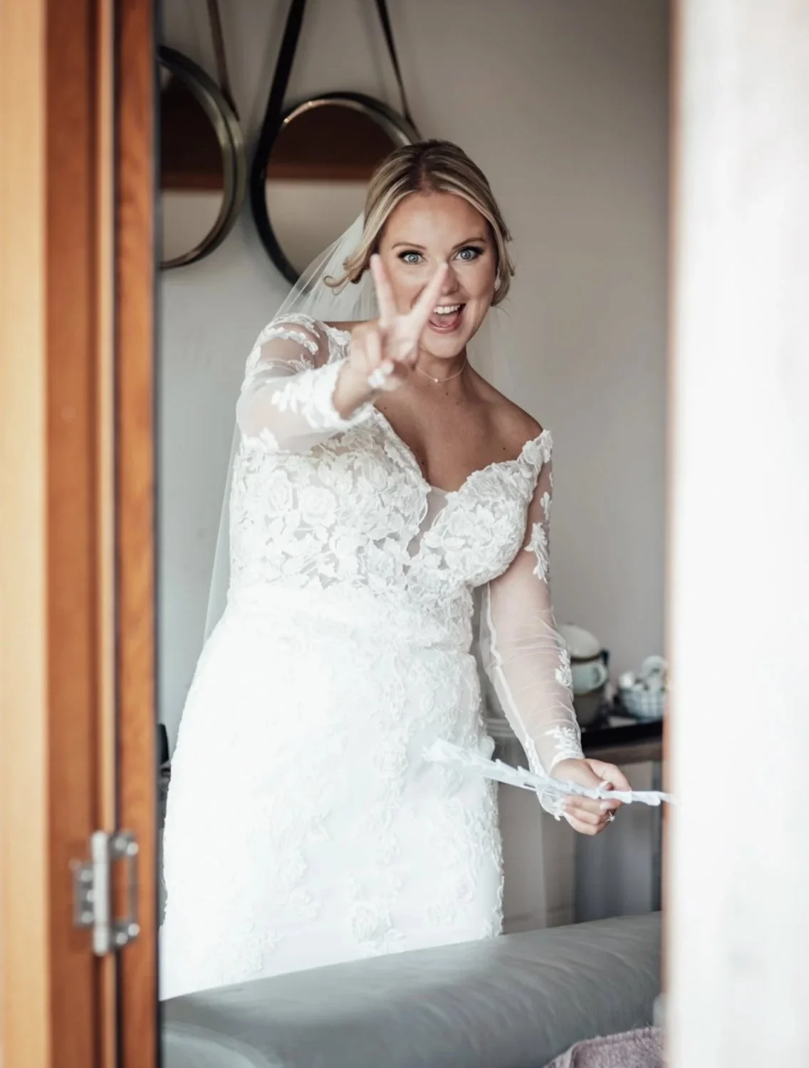 A bride in a white lace wedding dress taking a selfie in a mirror, making a peace sign with her hand.