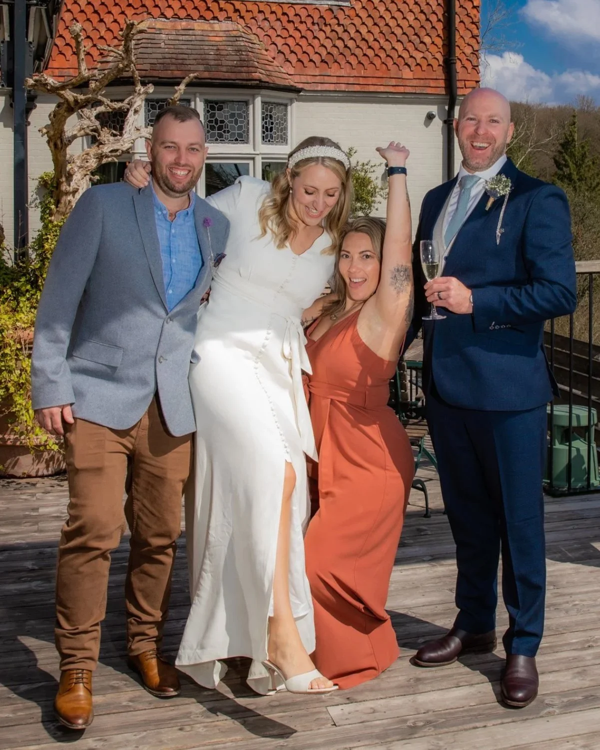 A group of five people celebrating outdoors in front of a house, including a bride in a white dress and a groom in a blue suit, with two other women and one man.