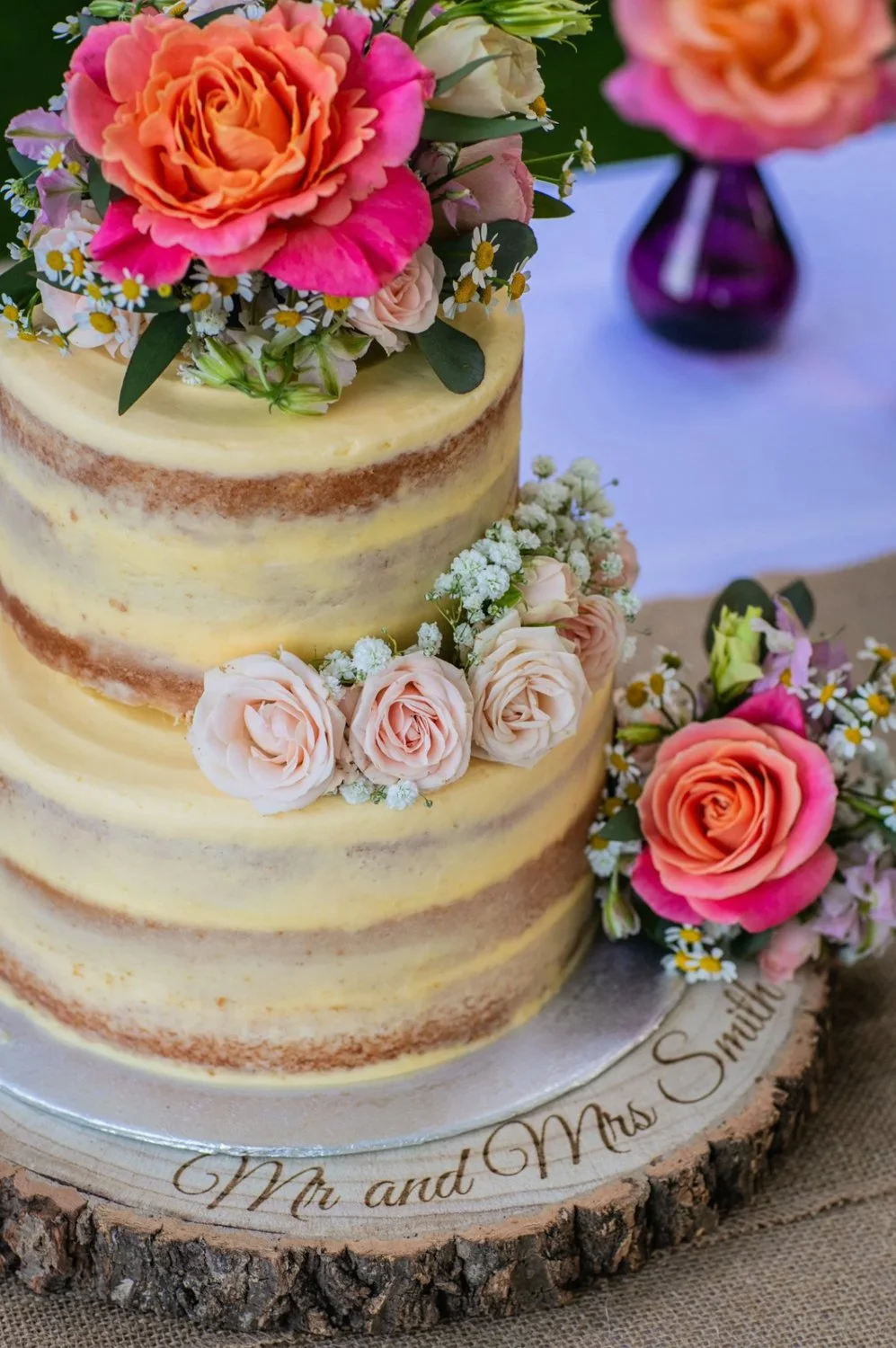 A three-layer wedding cake with a rustic, semi-naked style, decorated with pink, peach, and cream roses, small white daisies, and greenery, placed on a wooden slice base with 'Mr and Mrs Smith' written on it, surrounded by additional floral arrangeme