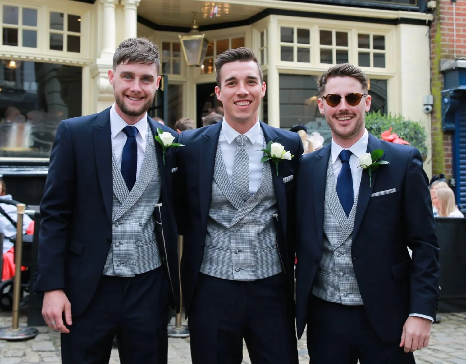 Three men in tuxedos with boutonnières smiling outdoors, standing close together.