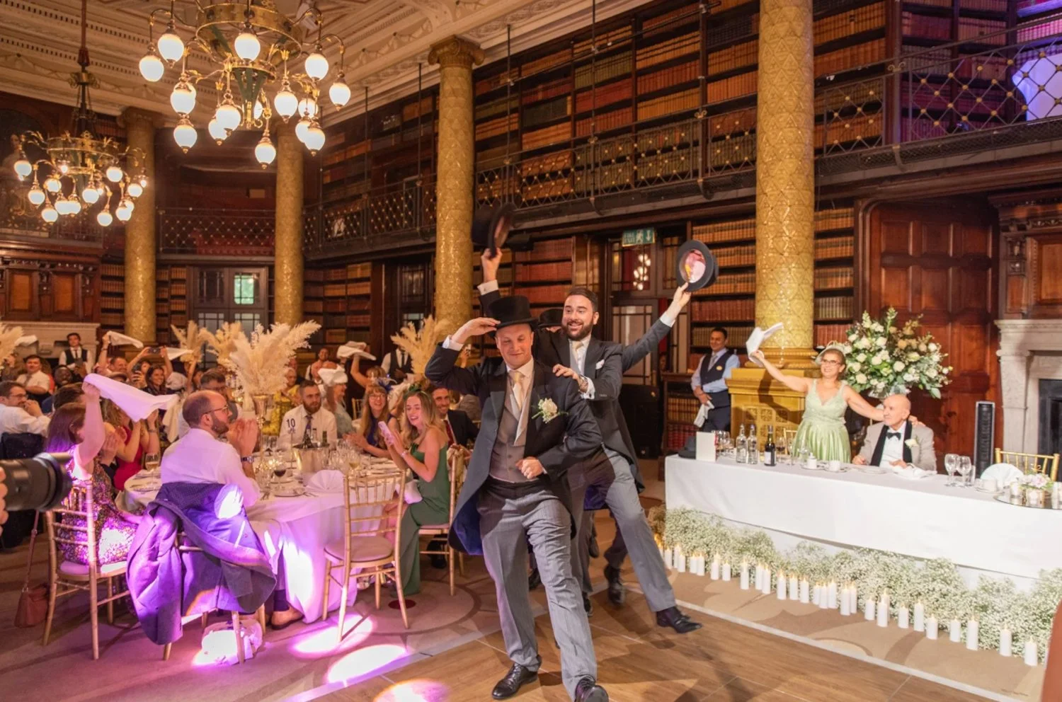 A wedding reception with guests seated at decorated tables, two performers in tuxedos, one wearing a top hat and the other holding a hat in the air, entertaining the crowd in a grand historic library-like venue with high ceilings, bookshelves, golden