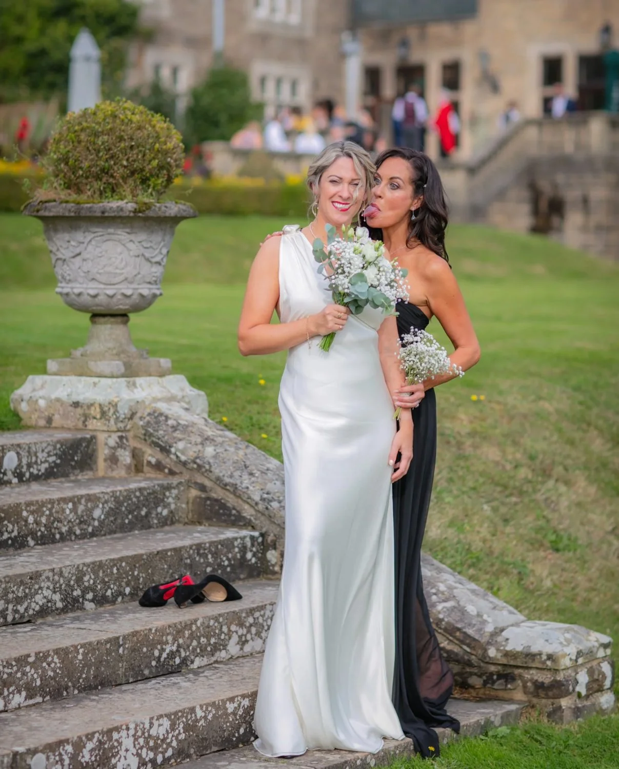 Two women in formal dresses, one in white and the other in black, standing on stone stairs outdoors. They are holding bouquets of white flowers. The woman in white is smiling and winking, while the woman in black is making a playful face. There are p