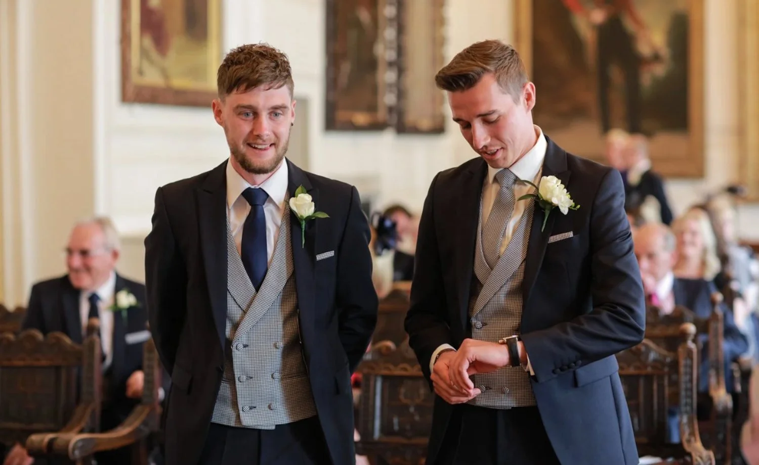 Two men in tuxedos with boutonnieres standing in a formal room, smiling and looking at a watch, with seated guests in the background.