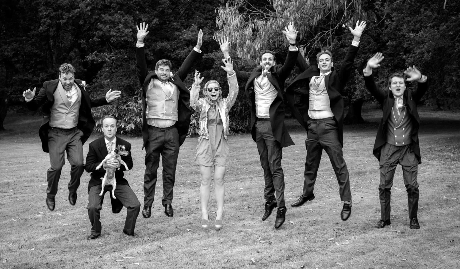 A group of seven people in formal attire jumping in the air outdoors, with trees and grass in the background.