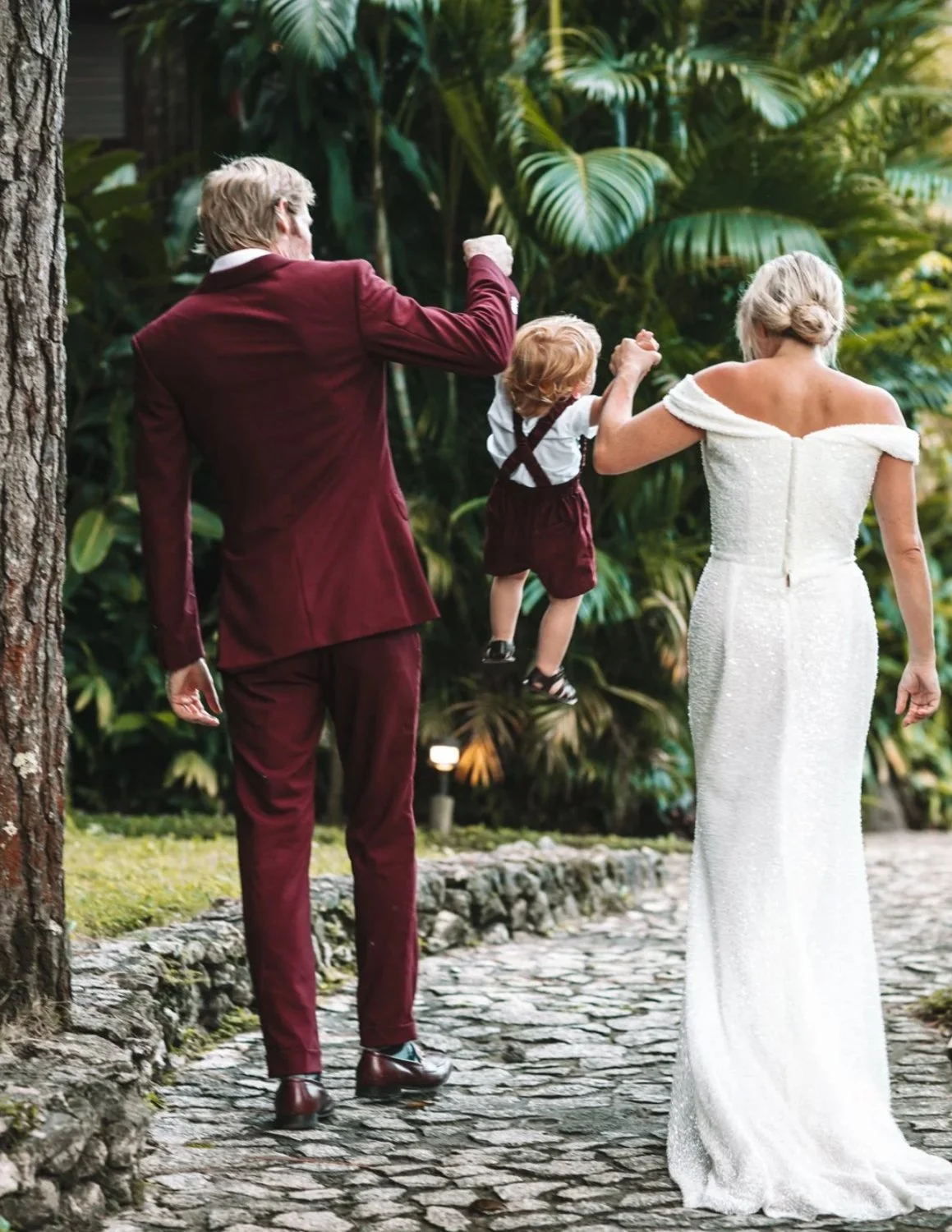 A couple, with the man dressed in a burgundy suit and the woman in a white gown, carry a small child dressed in suspenders and shorts along a stone path in a garden.
