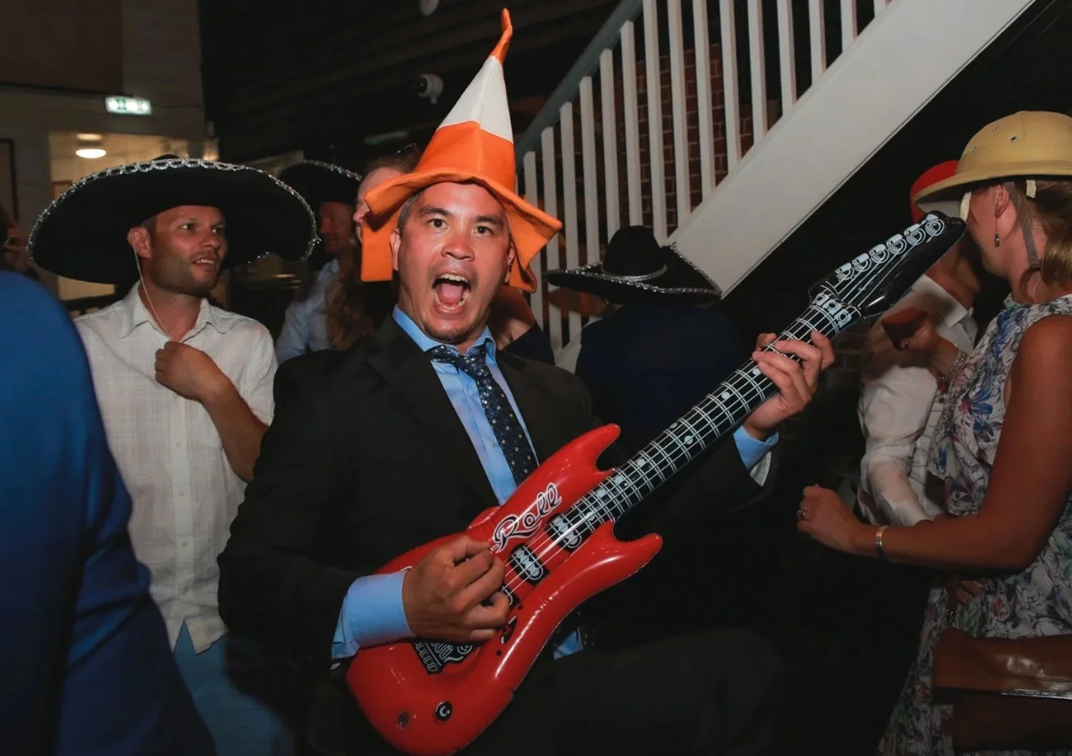 Man wearing a witch hat and suit, singing with an inflatable guitar at a party, surrounded by people in Mexican sombreros and hats.