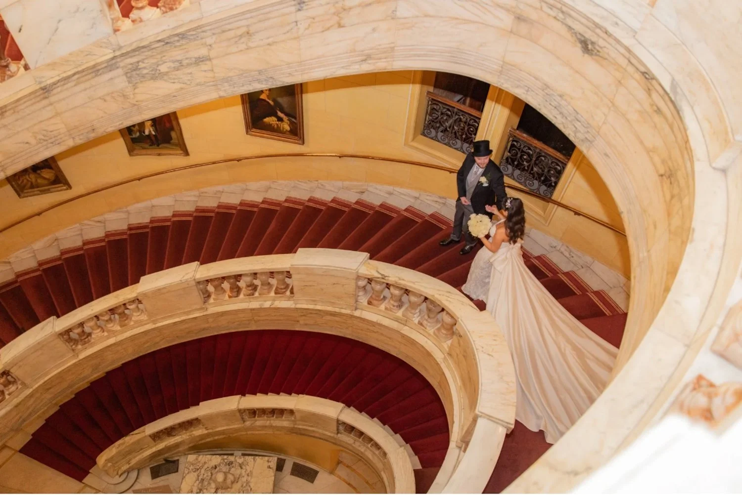 A bride and groom standing on a spiral staircase, with the bride holding a bouquet of flowers and wearing a wedding gown, and the groom dressed in a tuxedo with a top hat, seen from above in an elegant, historic building.