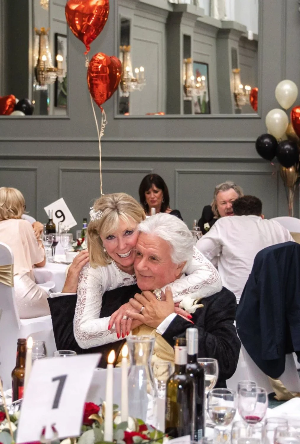 Two women at a formal celebration hugging and smiling, with balloons and guests seated at tables in the background.