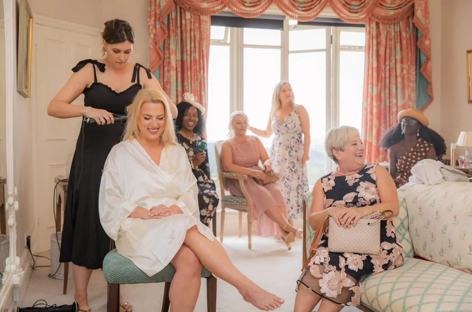 A group of women in a bright, elegant room preparing for a special occasion, with some getting hair and makeup done and others chatting and sitting on furniture.