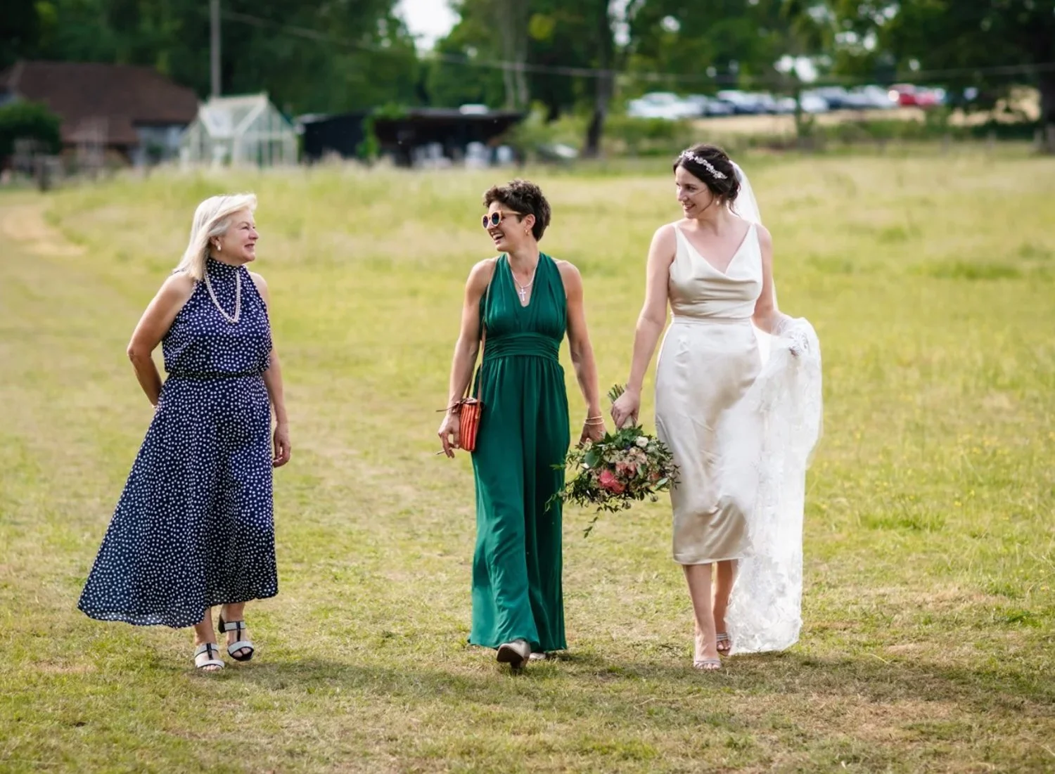 Four women walking on a grassy field, with the woman on the right wearing a white wedding dress and holding a bouquet, indicating a wedding scene; the other women are dressed in formal attire.