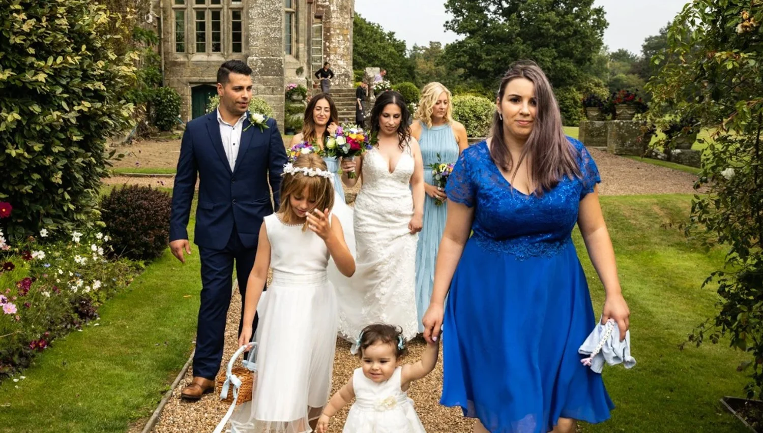 A group of people dressed in formal attire walking outside on a garden path, including women in dresses, some holding flowers, and children in white dresses, with a stone building in the background.