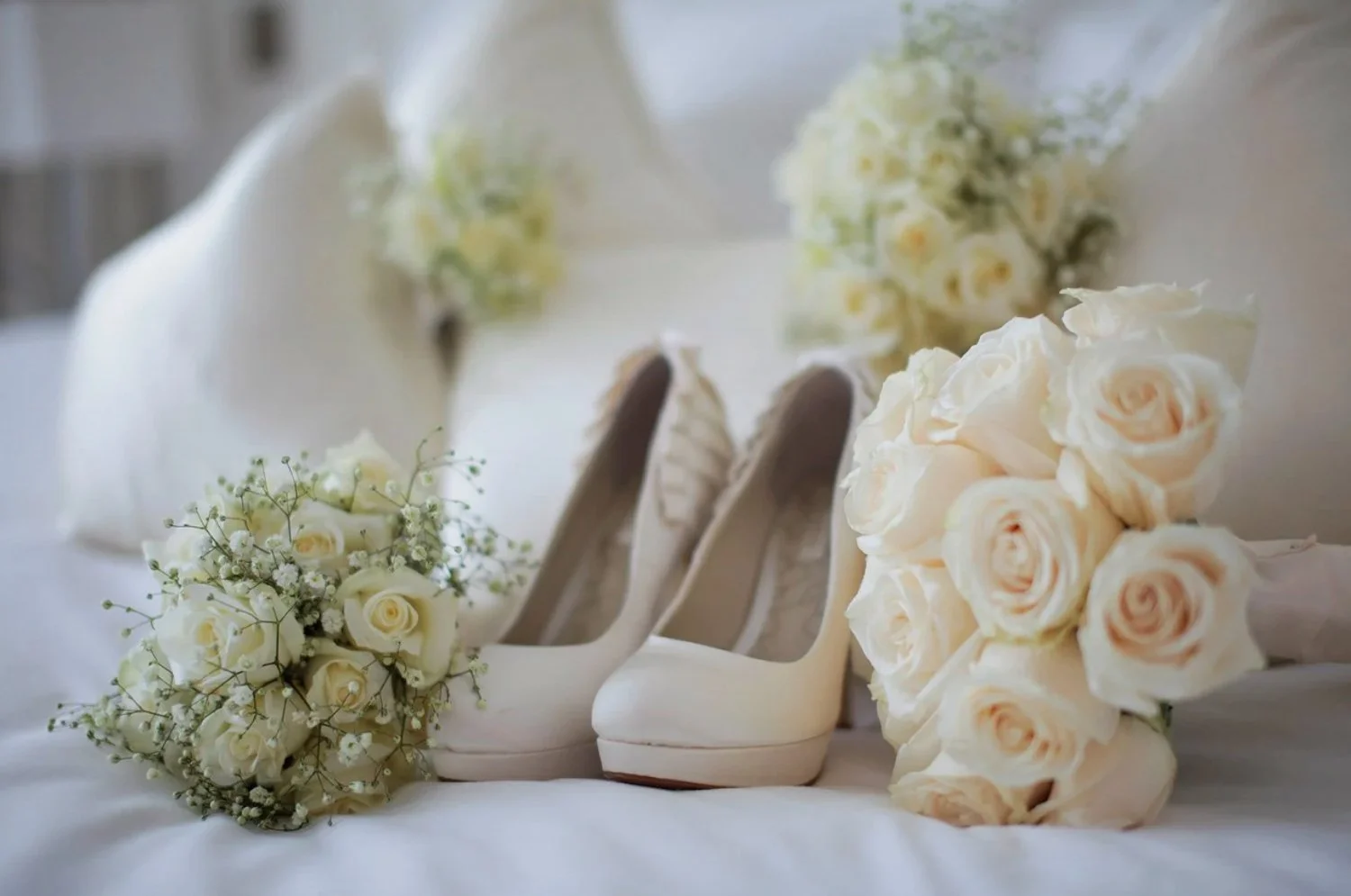 A pair of white high-heeled shoes, a bouquet of white roses, and another bouquet of white roses with baby's breath, arranged on a white surface, likely for a wedding.