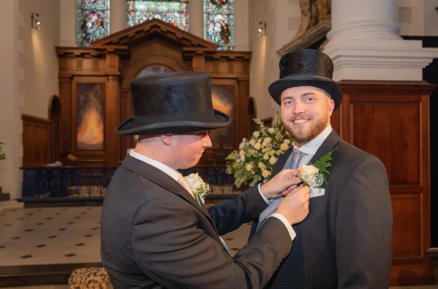 Two men in tuxedos and top hats at a wedding ceremony, one helping the other pin a boutonniere on his lapel inside a church with stained glass windows and floral arrangements in the background.