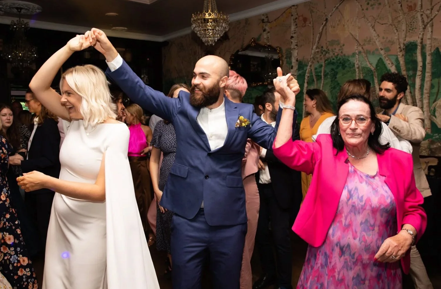 People dancing at a wedding reception, with a man in a blue suit holding hands with a woman in a white dress and a woman in a pink blazer smiling and holding hands in the foreground, with other guests dancing in the background.