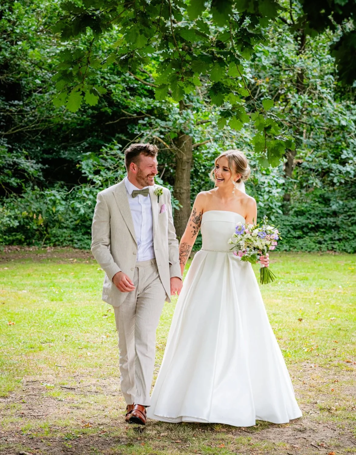 A bride and groom walking hand in hand outdoors, smiling and looking at each other, surrounded by green trees and grass, during their wedding ceremony.