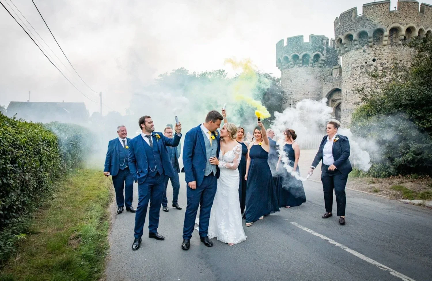 Wedding party on a road in front of a castle, with some members holding smoke bombs and taking photos.
