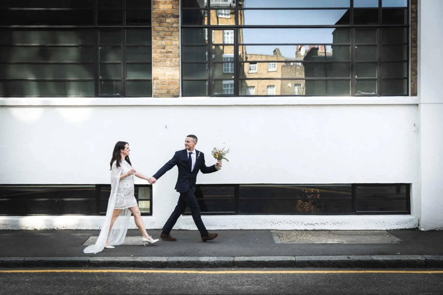 A bride and groom holding hands and walking on a city sidewalk, with the groom holding a bouquet of flowers, outside a building with large window panes.