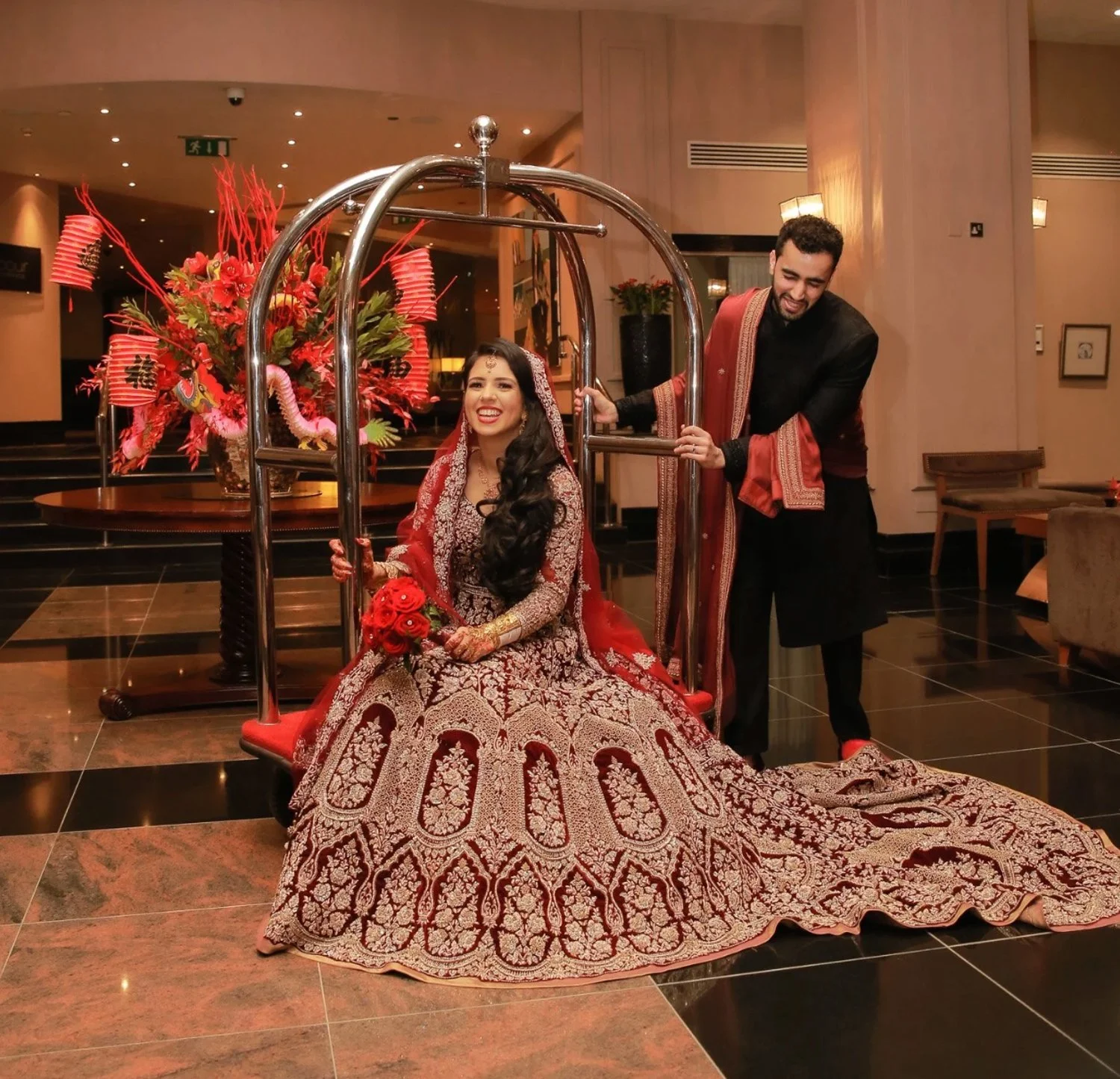 A bride in a traditional ornate red and gold wedding dress, sitting in a luggage cart with a bouquet of red roses, smiling. A groom dressed in black with a red stole, pushing the cart, also smiling. Behind them is a large floral arrangement with red
