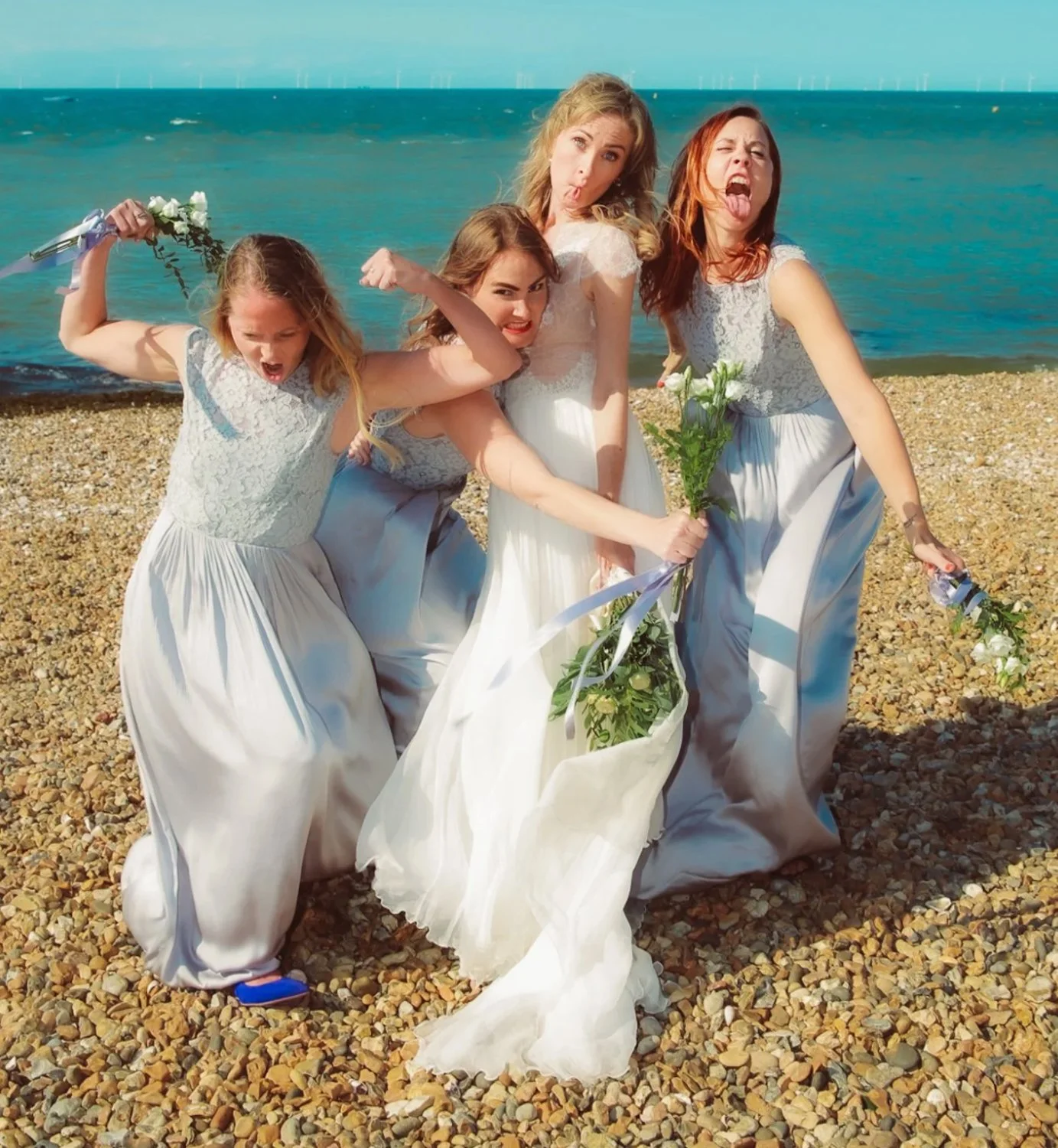 Four women in white dresses at the beach, making silly faces and posing playfully with flowers.