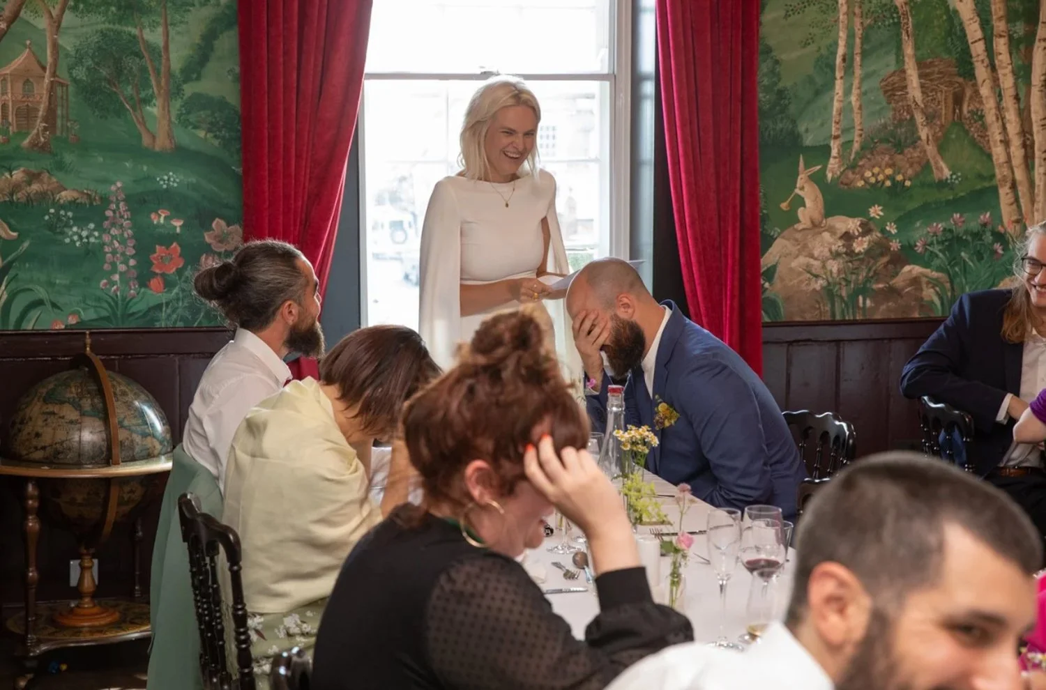 A woman in white standing and smiling at a wedding reception, with guests sitting at a table in front of her, some covering their faces and appearing emotional, inside a room with red curtains and a decorative mural on the walls.