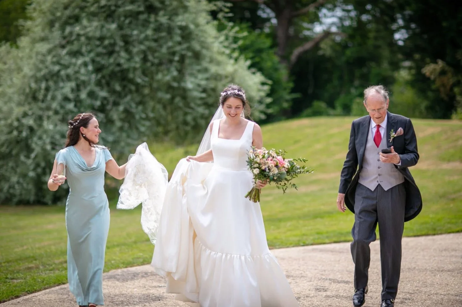 A bride in a white wedding dress holding a bouquet, walking with a woman in a light blue dress and a man in a suit, outdoors with green trees in the background.