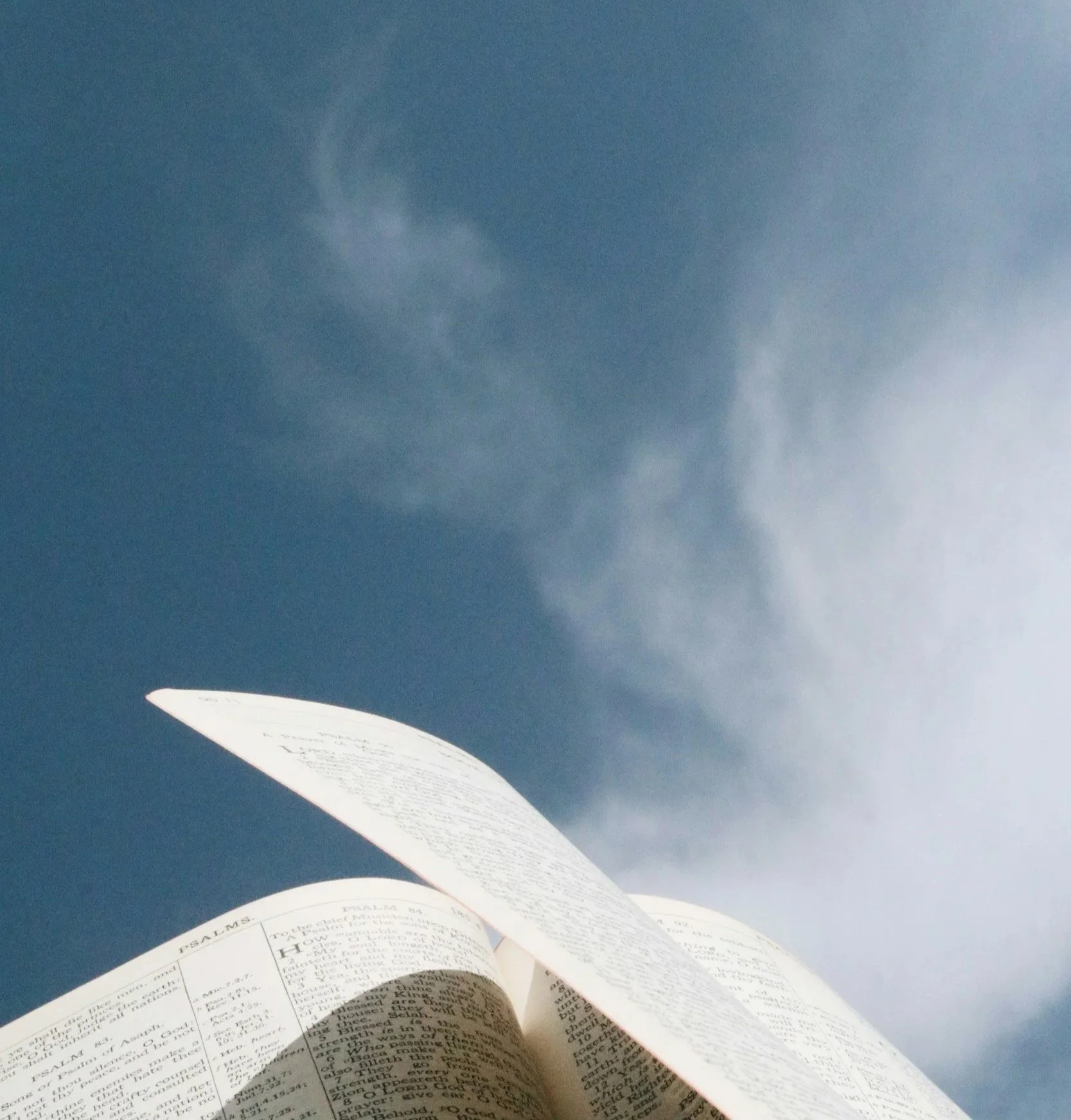 Open Bible with pages flipping against a sky background with wispy clouds.