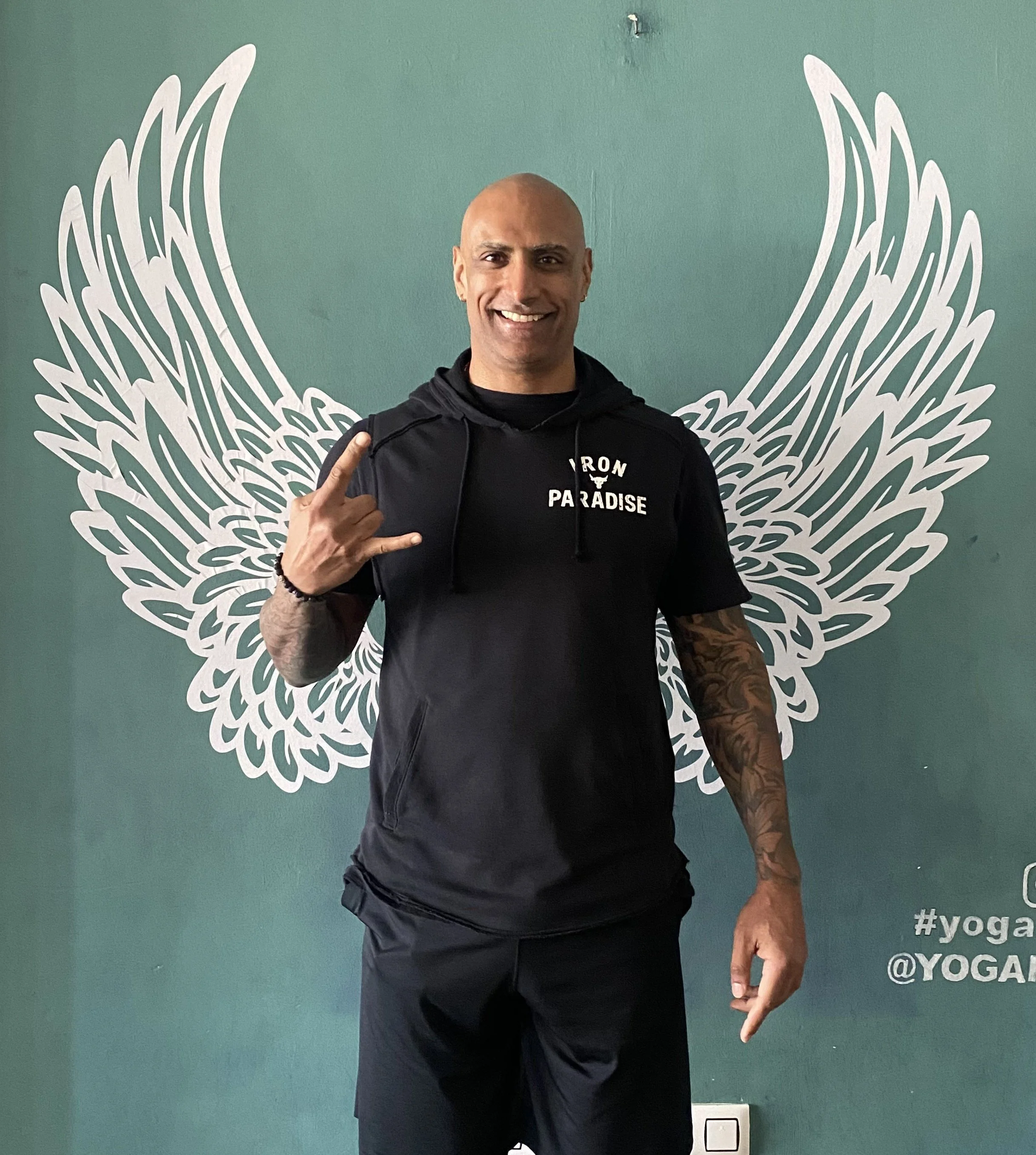 Man smiling, making a rock sign with his right hand, standing in front of a wall with angel wings design painted behind him, wearing a black hoodie and black shorts.