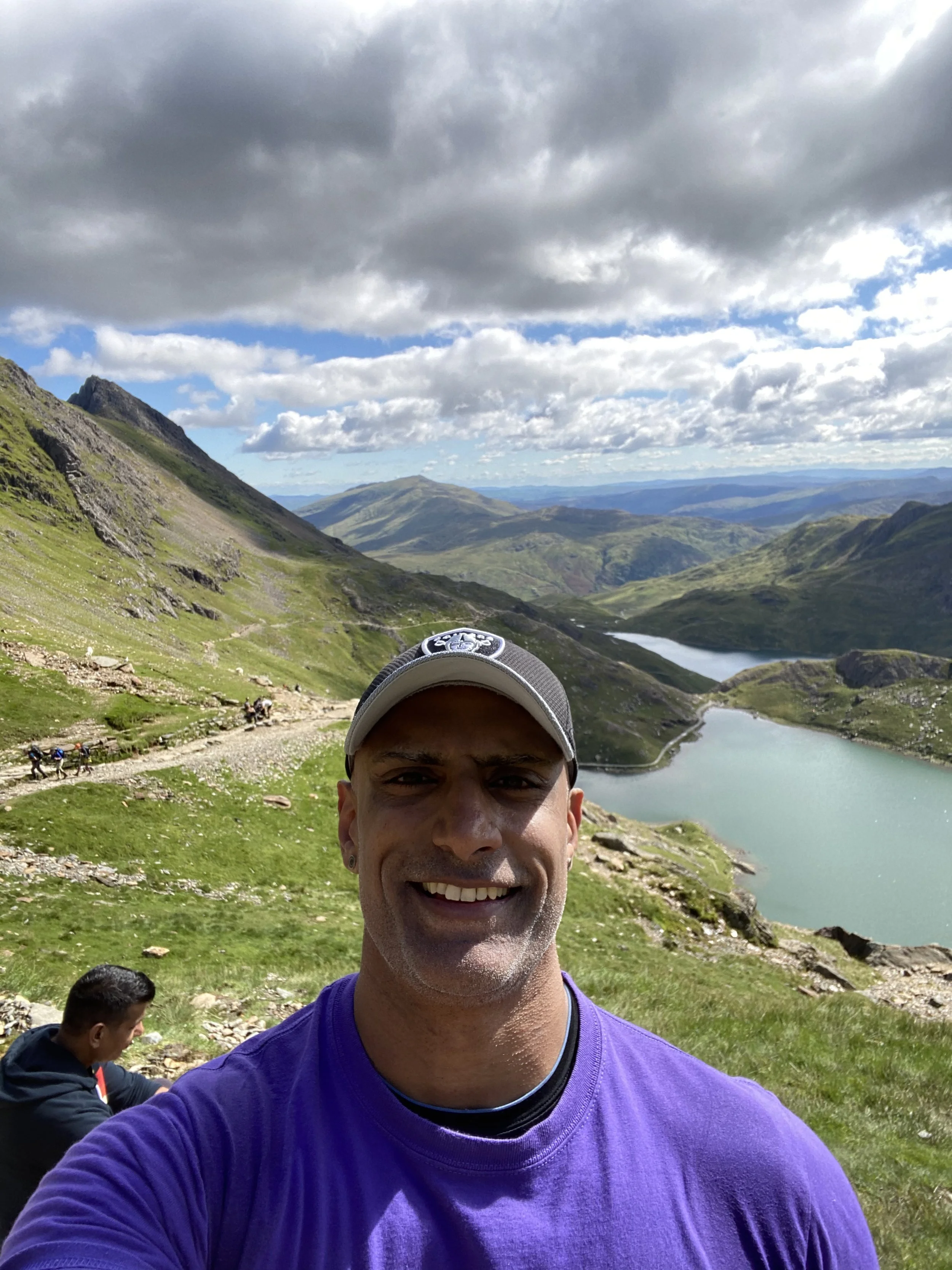 A smiling man wearing a baseball cap and purple shirt takes a selfie in a mountainous landscape with green hills, a lake, and a partly cloudy sky.