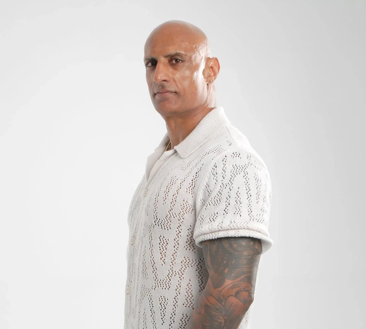 A bald man with light skin and a serious expression, wearing a short-sleeved white button-up shirt with a patterned weave, standing against a plain white background.