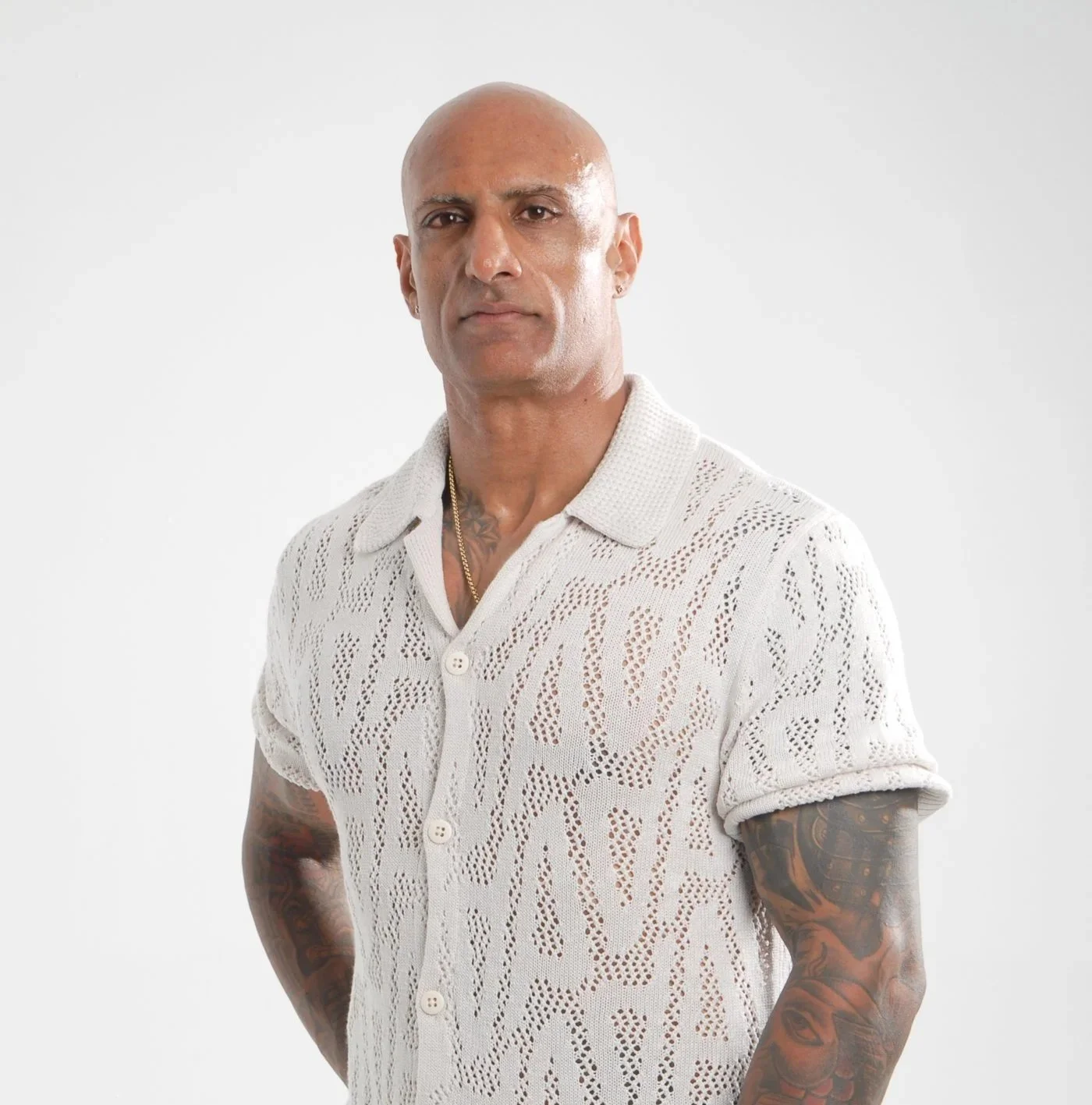 Bald man with tattoos wearing a white button-up shirt with a pattern, standing against a plain white background.