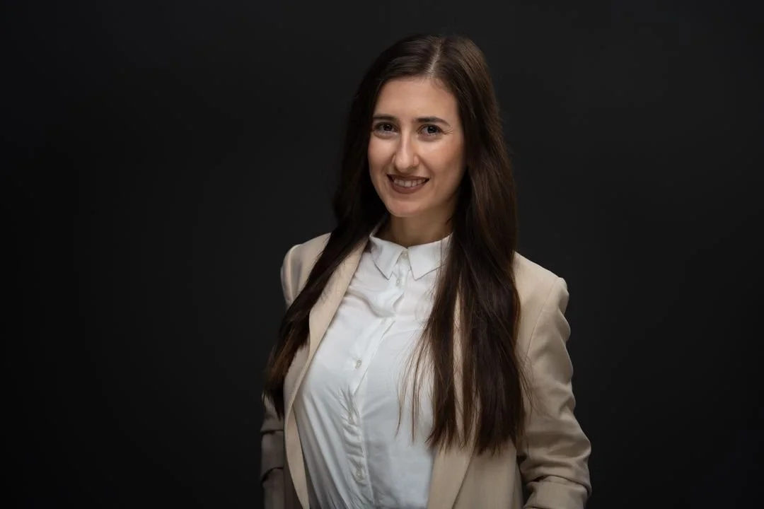 A woman with long dark hair smiling at the camera against a dark background, wearing a light-colored blazer over a white button-up shirt.