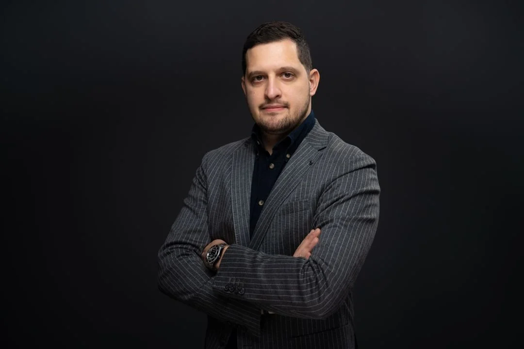 A man in a gray pinstripe suit with a black shirt, standing with arms crossed against a dark background.