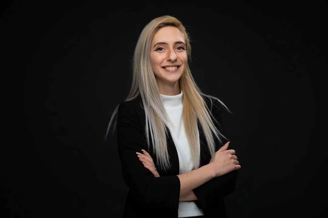 A smiling blonde woman with long hair, wearing a black blazer and white top, standing against a black background with arms crossed.