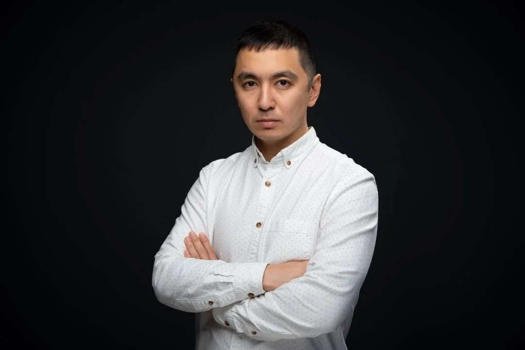 A man with short black hair and light skin, wearing a white button-up shirt, stands with arms crossed against a dark background.