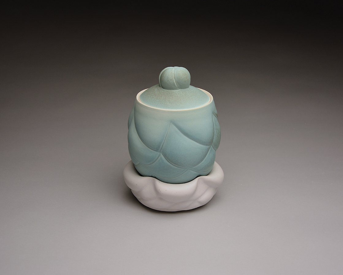 A decorative ceramic container in the shape of an artichoke with a white base and light green top, placed on a gray surface with a dark gray background.