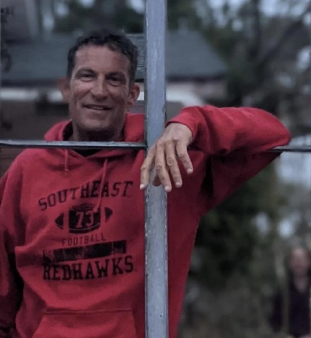 A man with short dark hair wearing a red sweatshirt with 'SOUTHEAST FOOTBALL REDHAWKS' printed on it, smiling and resting one arm on a metal structure outdoors.