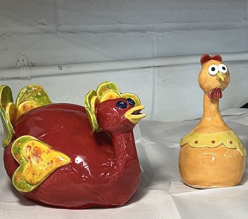 Lizette Terral Pottery Artist Colorful ceramic sculptures of a turkey and a chicken, with the turkey having a round red body and green and yellow tail feathers, and the chicken having a yellow-orange body with a red comb and wattles.
