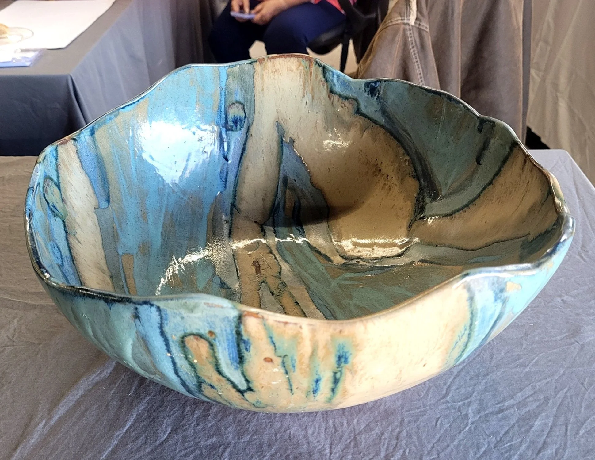 A colorful ceramic art bowl with a wavy rim, featuring blue, beige, and black glaze patterns, placed on a table.