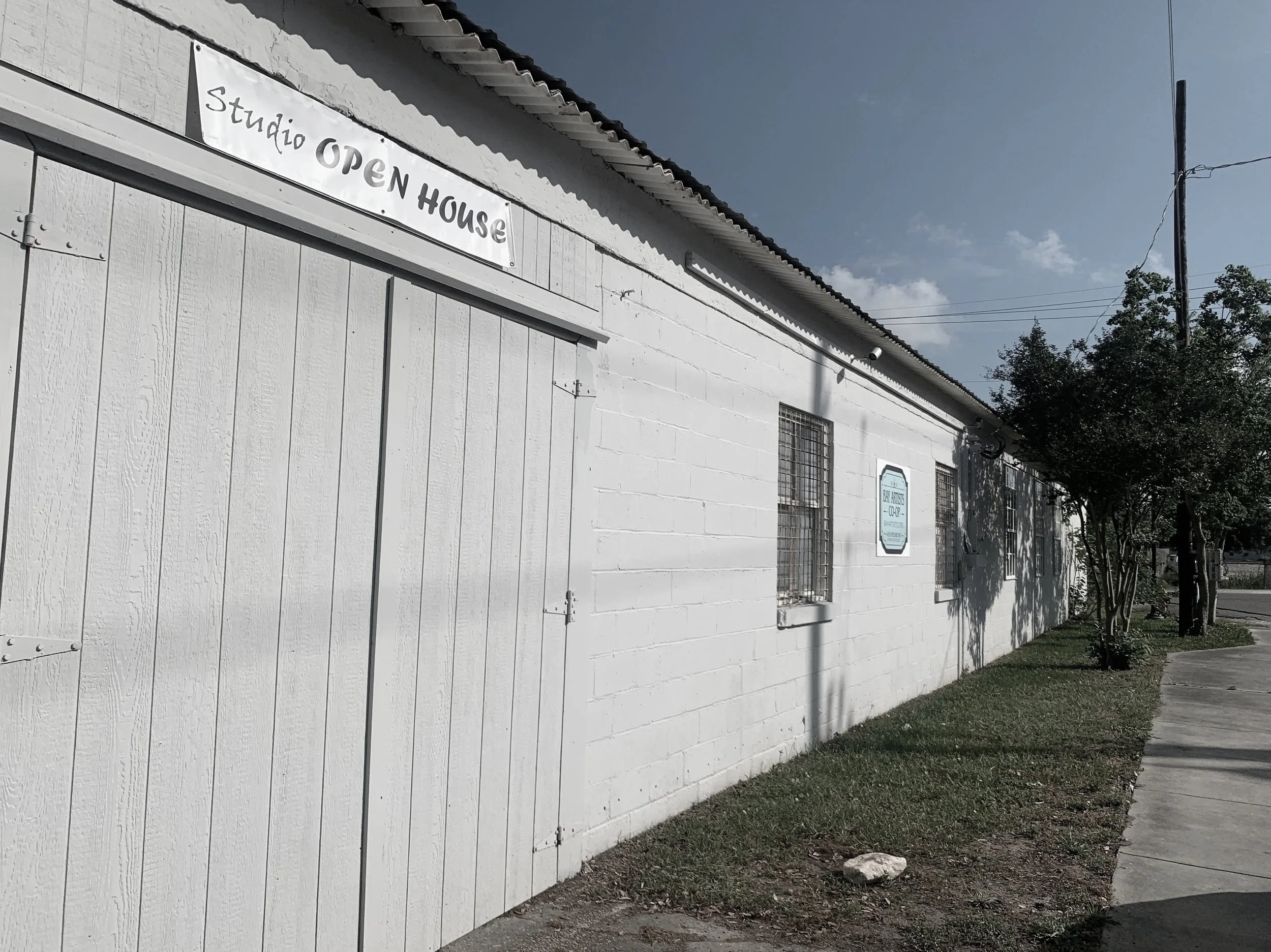 White building with a sign that reads 'Studio OPEN HOUSE', barred windows, trees along the sidewalk, and utility pole against a partly cloudy sky.