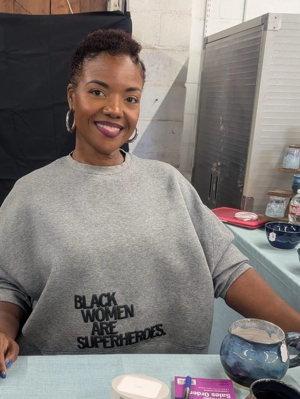 Thomika Segar Artist Pottery. Smiling woman sitting at a table wearing a gray sweatshirt that says 'Black Women Are Superheroes' with a black background and kitchen items on the side.