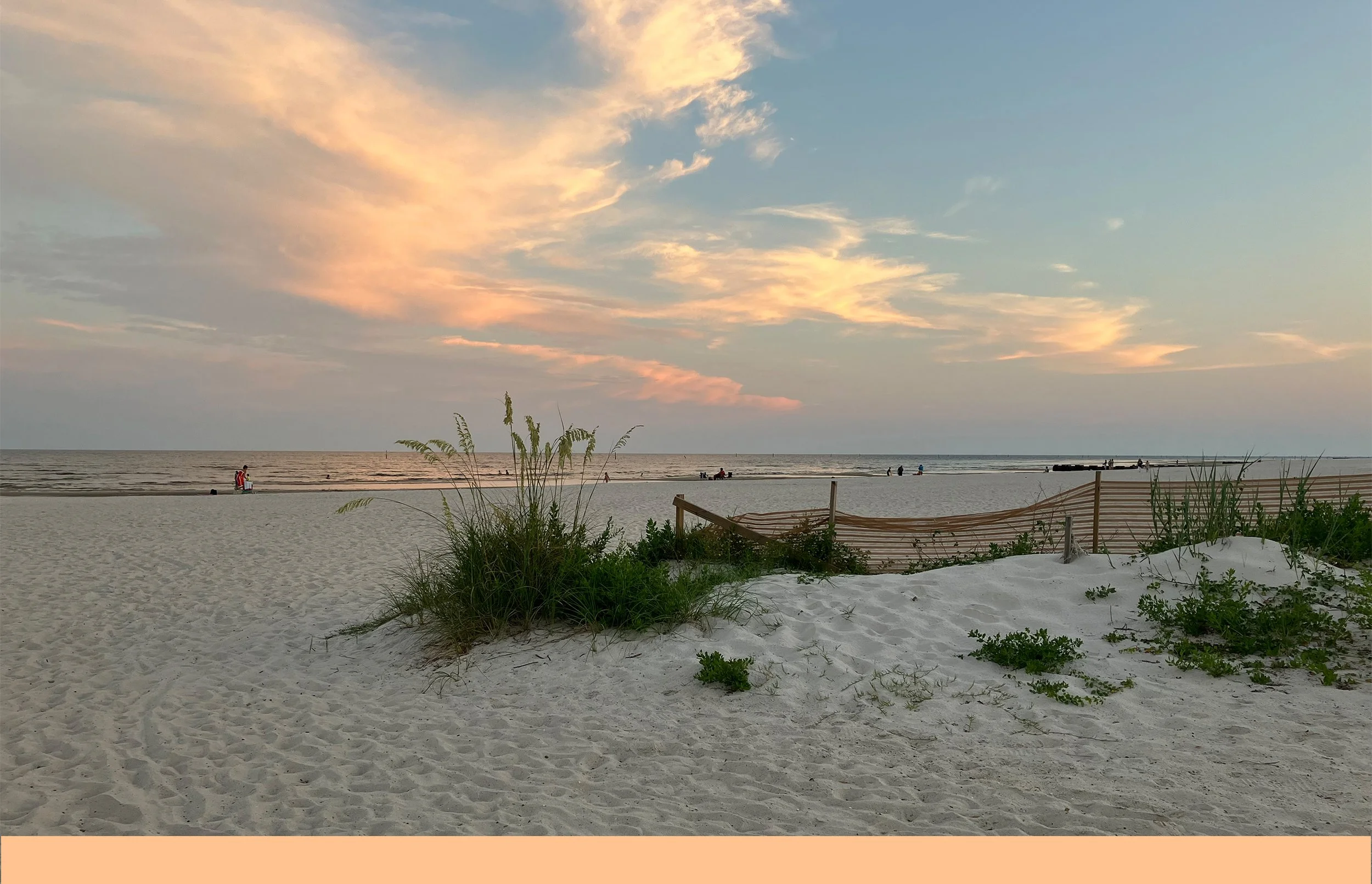 A peaceful beach scene at sunset with soft pink and orange clouds in the sky, sandy shore with green plants, a wooden barrier, and people walking along the shoreline.