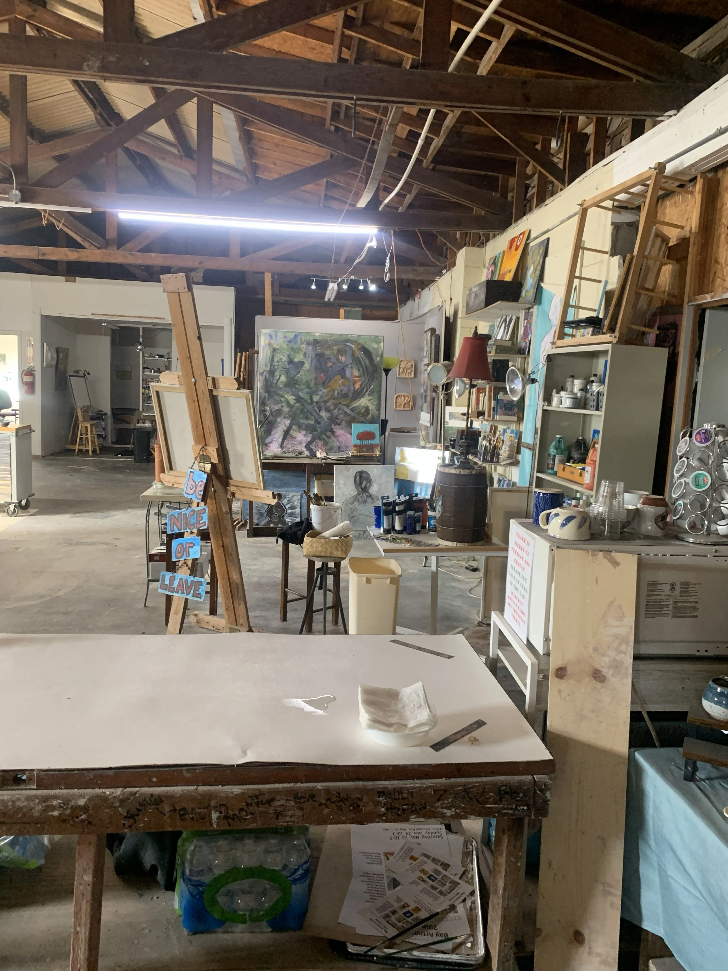 Interior of an art studio with paintings, art supplies, and a sign that reads 'Be Nice or Leave' on an easel. The space has exposed wooden beams and a cluttered, creative atmosphere.