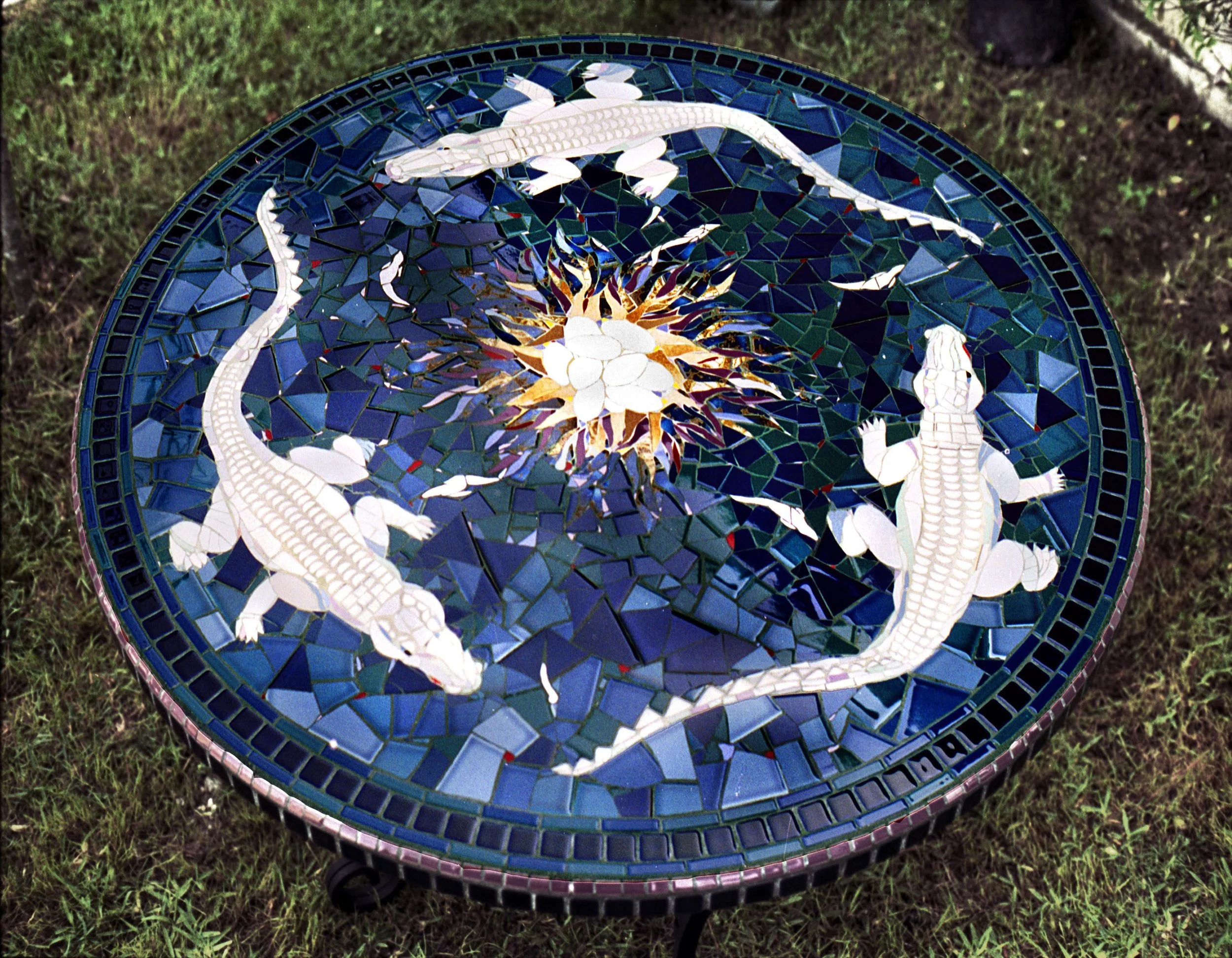 Elizabeth Veglia Artist Jewelry Maker Mosaic Artist A circular mosaic table featuring two alligator figures, a central sun with radiating pointed shapes, and a dark blue tiled background, set on grass.