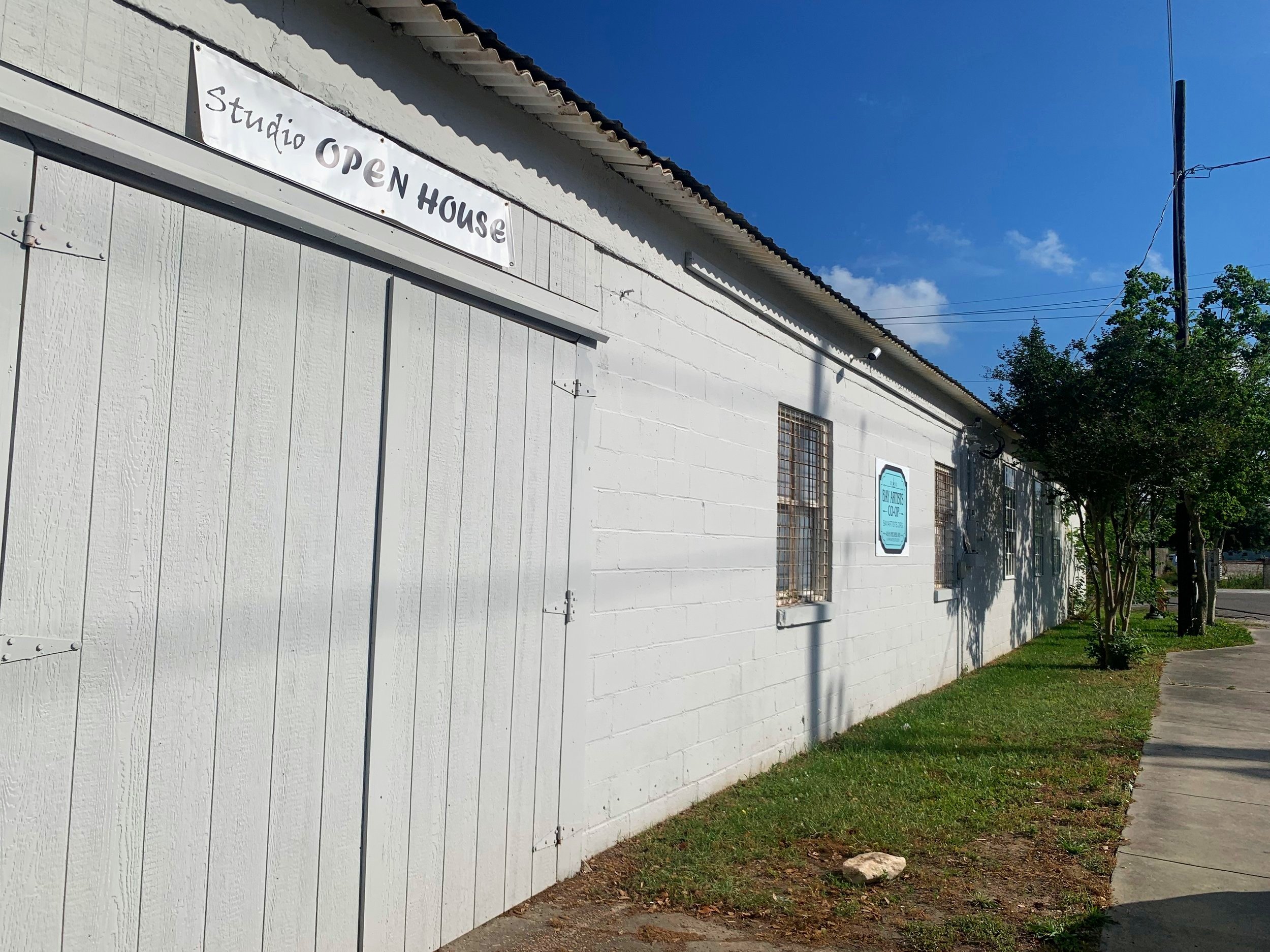 Exterior of a white building with a sign that reads "studio OPEN HOUSE" and a blue sign that says "Barber Studio & Barbershop." There are trees and a sidewalk in front on a sunny day with a clear blue sky.