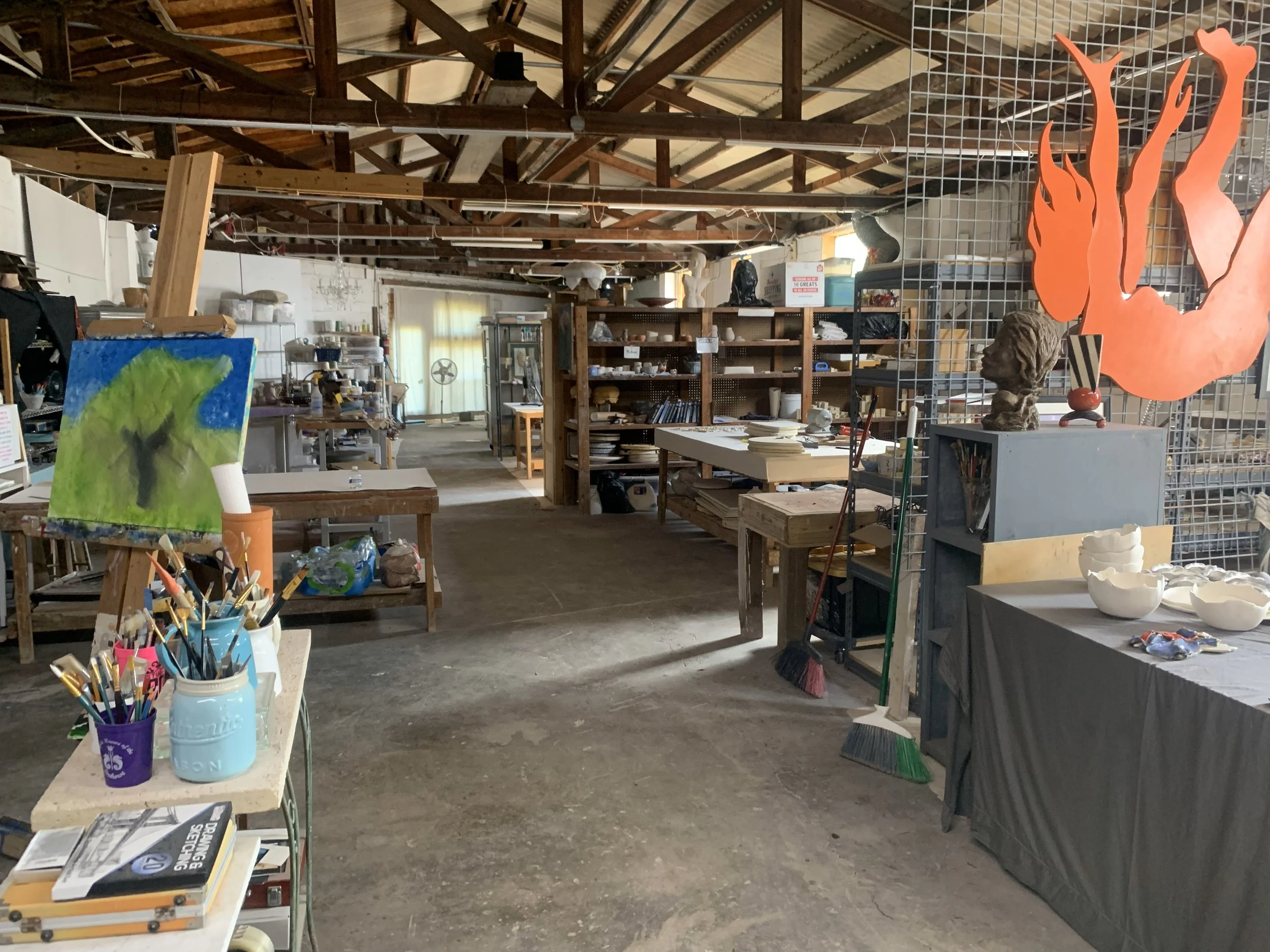 An art studio with shelves of supplies, tables with various art projects, painting on an easel, and art sculptures, including a large orange flame-shaped piece.