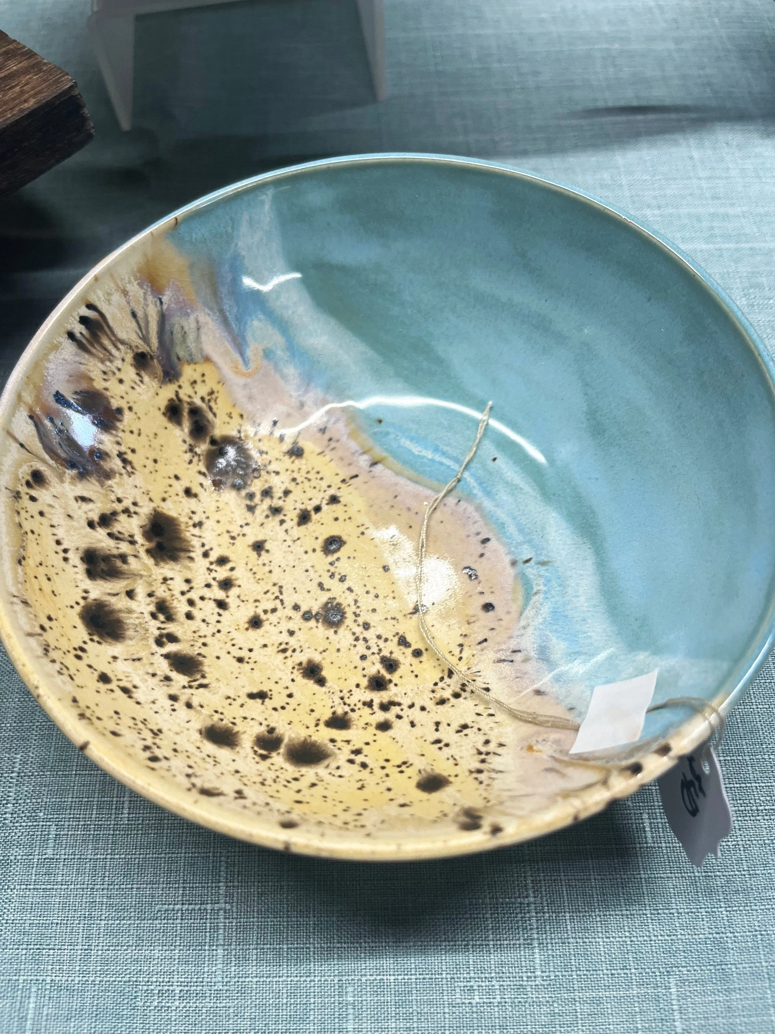 Thomika Segar Artist Pottery. ceramic bowl with a light blue interior and yellow exterior, containing dark brown coffee stains and a stray hair, placed on a greyish-green textured tablecloth.