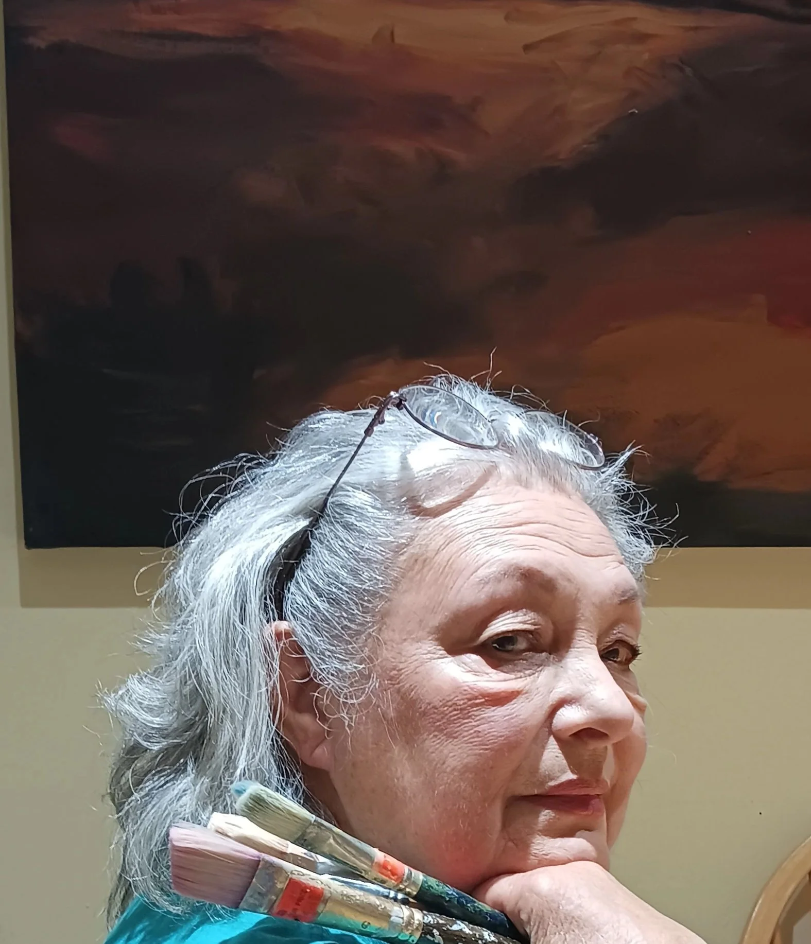 An elderly woman with gray hair, wearing glasses on her head, holds paintbrushes and looks to the side. A dark abstract painting is in the background.