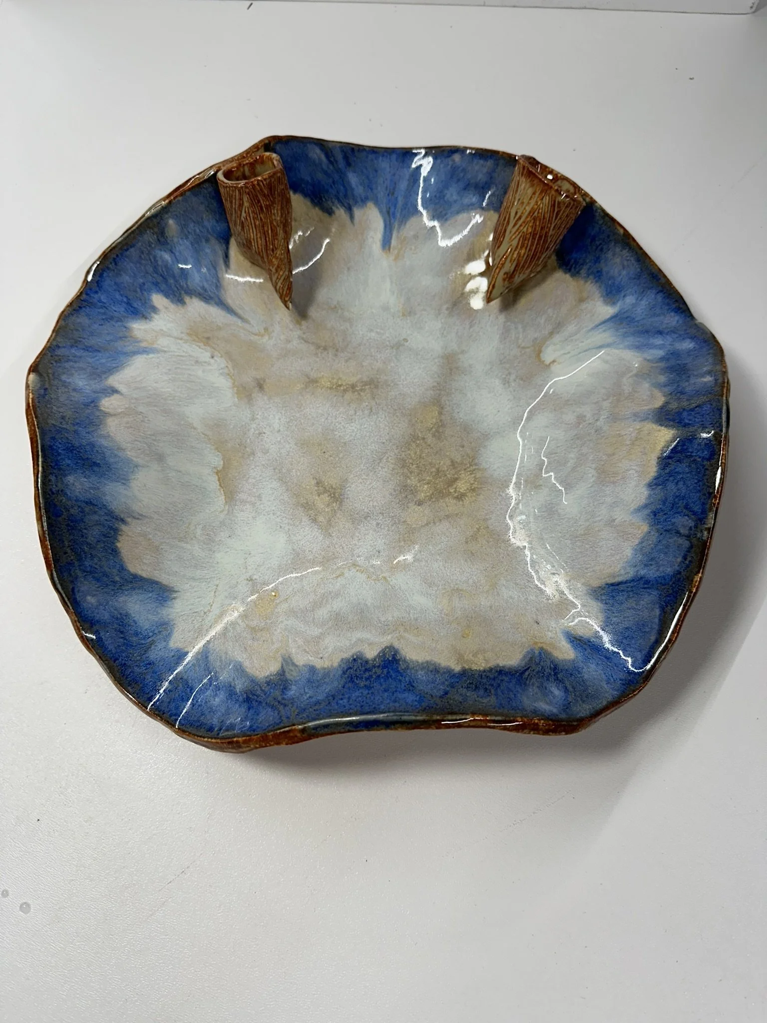 Decorative ceramic bowl with blue, white, and brown glaze, featuring two brown textured handles, placed on a white surface.