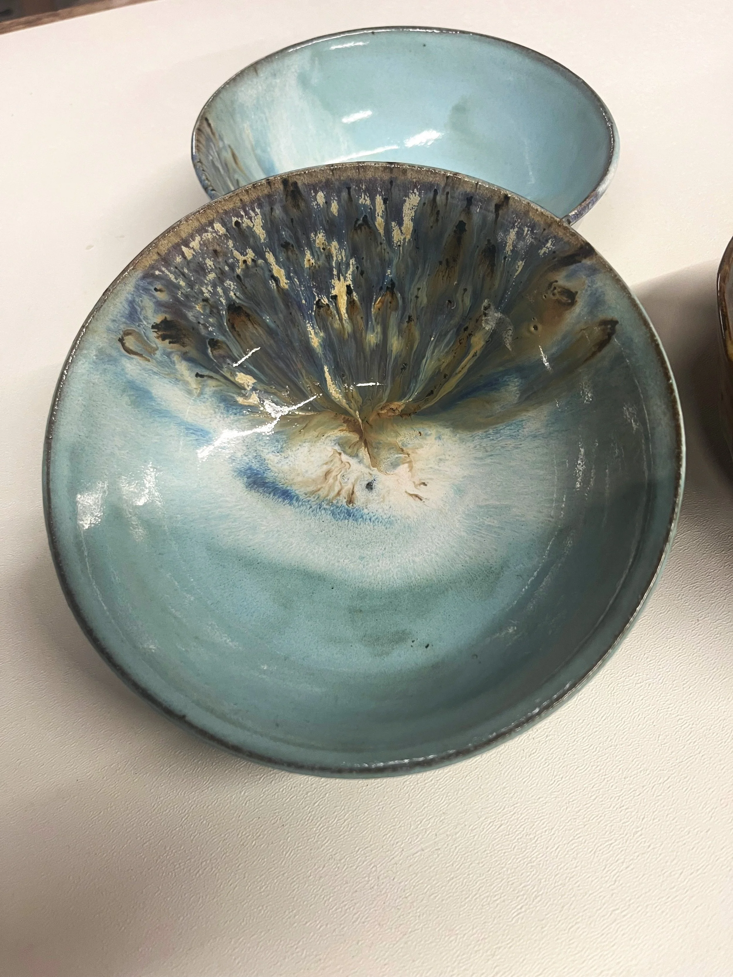 Thomika Segar Artist Pottery. Two ceramic bowls with blue and brown glaze, one larger and in front, the other smaller and behind, on a white surface.
