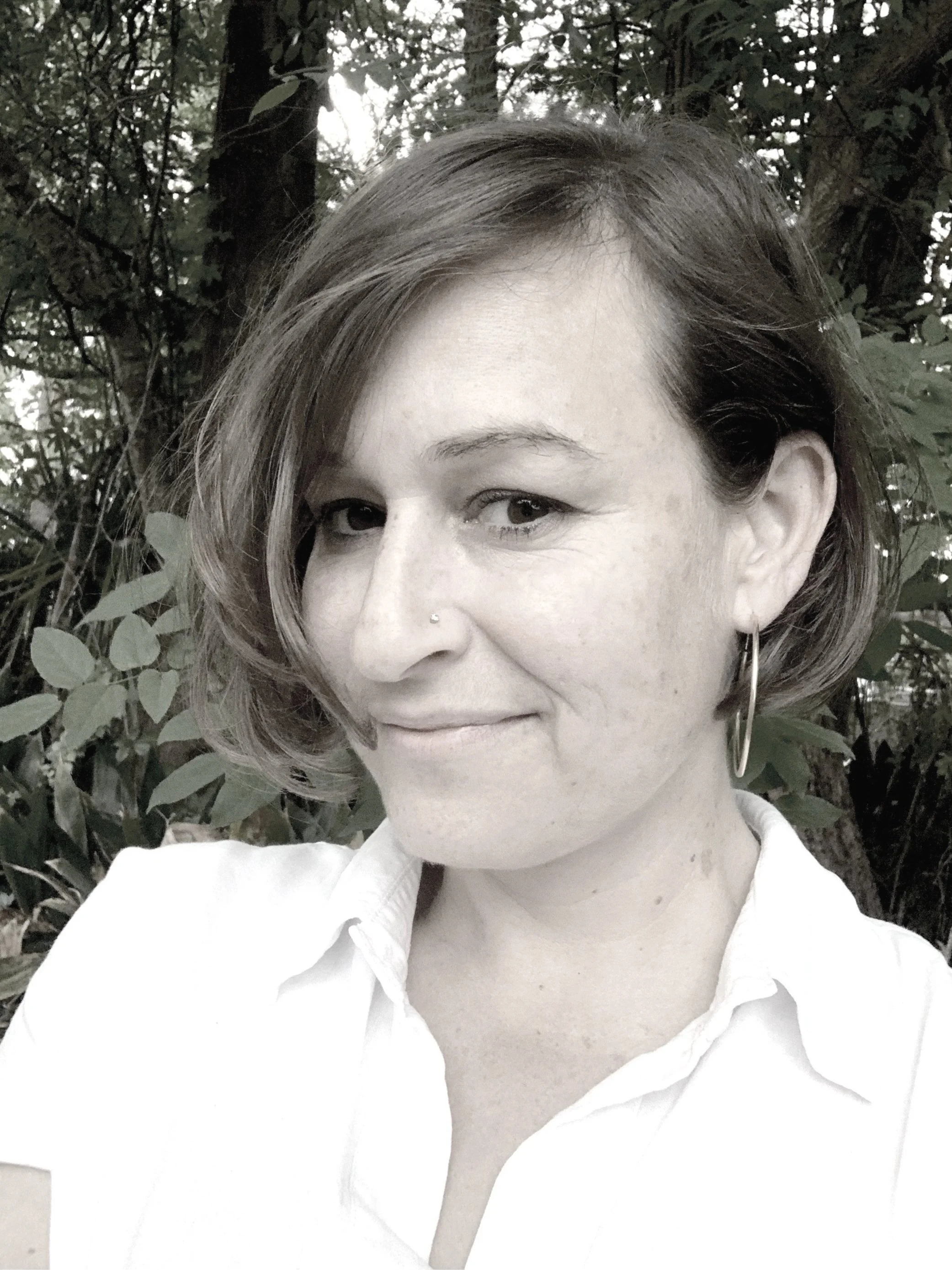 Woman with short wavy hair, wearing hoop earrings and a nose piercing, in a white collared shirt, outdoors with greenery in the background.